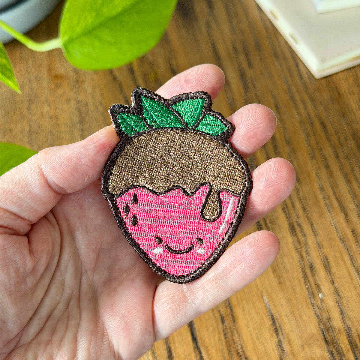 A hand holding a cute embroidered strawberry patch with a smiling face, pink body, brown top, and green leaves, set against a wooden table background.