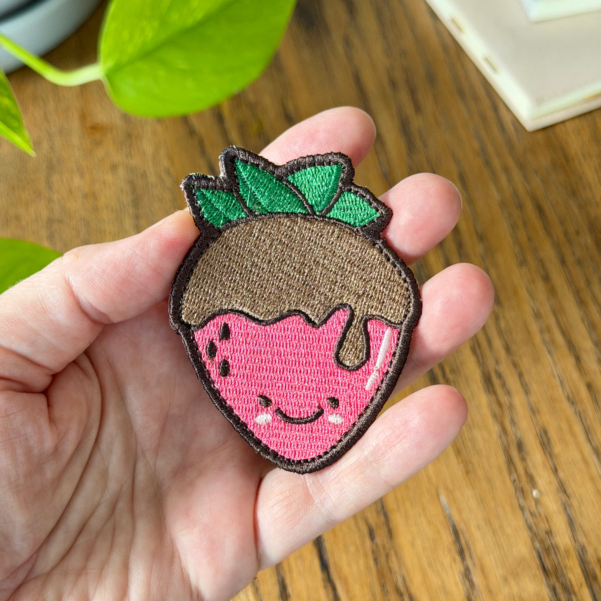 A hand holding a cute embroidered strawberry patch with a smiling face, pink body, brown top, and green leaves, set against a wooden table background.